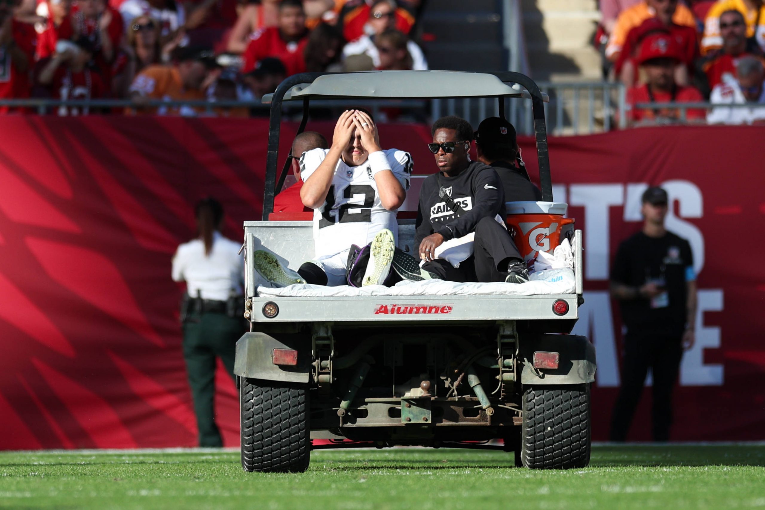 Ein Footballspieler, Raiders-Quarterback Aidan O'Connell, sitzt in einer weiß-schwarzen Uniform auf der Rückseite eines Wagens und bedeckt sein Gesicht mit den Händen, wahrscheinlich aufgrund einer Knieverletzung. Neben ihm steht eine weitere Person in Schwarz. Sie befinden sich auf einem Footballfeld mit einer Menschenmenge im Hintergrund, von denen einige rot gekleidet sind. Diese Beschreibung wurde mit der FootballR KI automatisch generiert.