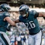 Zwei Footballspieler in Philadelphia Eagles-Uniformen jubeln auf dem Spielfeld. Der Spieler links, Nummer 81, streckt seinen Arm in Richtung des Spielers rechts, Nummer 26 - Saquon Barkley, aus. Sie scheinen einen jubelnden Moment zu erleben, im Hintergrund ist die Stadionmenge verschwommen. Diese Beschreibung wurde mit der FootballR KI automatisch generiert.