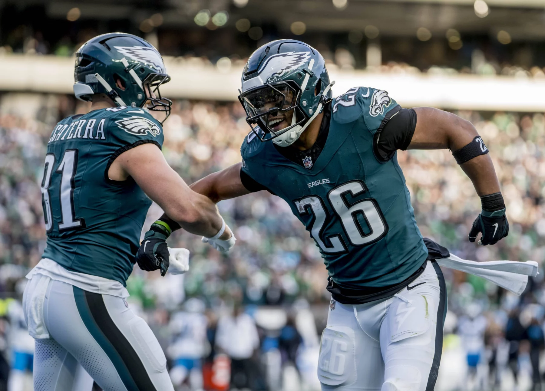 Zwei Footballspieler in Philadelphia Eagles-Uniformen jubeln auf dem Spielfeld. Der Spieler links, Nummer 81, streckt seinen Arm in Richtung des Spielers rechts, Nummer 26 - Saquon Barkley, aus. Sie scheinen einen jubelnden Moment zu erleben, im Hintergrund ist die Stadionmenge verschwommen. Diese Beschreibung wurde mit der FootballR KI automatisch generiert.
