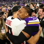 Zwei American-Football-Spieler umarmen sich nach einem Spiel herzlich auf dem Feld. Der Spieler im weißen Trikot mit roten Akzenten und der Aufschrift „COUSINS“ ist kein anderer als Kirk Cousins vom Team Minnesota. Der Spieler in Lila lächelt breit, während Fotografen und Medienpersonal ihn umringen und den Moment festhalten. Diese Beschreibung wurde mit der FootballR KI automatisch generiert.