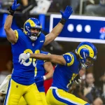 Zwei Footballspieler in blau-gelben Los Angeles Rams Trikots jubeln triumphierend auf dem Spielfeld. Ein Spieler hat die Arme erhoben, während der andere sich nach vorne beugt. Beide strahlen Begeisterung aus. Im Hintergrund ist die Menge unscharf zu sehen, über ihnen leuchtet eine leuchtende Anzeigetafel. Diese Beschreibung wurde mit der FootballR KI automatisch generiert.