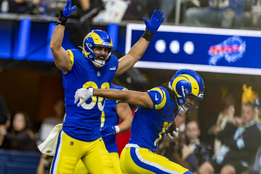 Zwei Footballspieler in blau-gelben Los Angeles Rams Trikots jubeln triumphierend auf dem Spielfeld. Ein Spieler hat die Arme erhoben, während der andere sich nach vorne beugt. Beide strahlen Begeisterung aus. Im Hintergrund ist die Menge unscharf zu sehen, über ihnen leuchtet eine leuchtende Anzeigetafel. Diese Beschreibung wurde mit der FootballR KI automatisch generiert.