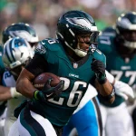 Ein Footballspieler in einer grünen Philadelphia Eagles-Uniform, Nummer 26, läuft mit dem Ball, umgeben von Teamkollegen und Gegnern. Im Hintergrund sind ein Teamkollege in einer ähnlichen Uniform und ein Gegner in einer blau-weißen Uniform zu sehen, zusammen mit verschwommenen Zuschauern im Stadion. Diese Beschreibung wurde mit der FootballR KI automatisch generiert.