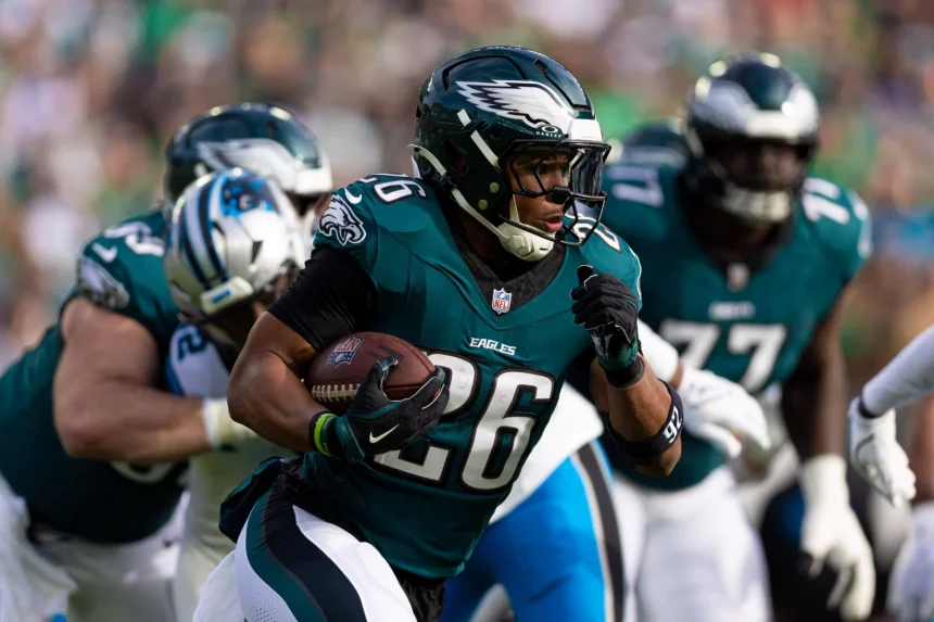 Ein Footballspieler in einer grünen Philadelphia Eagles-Uniform, Nummer 26, läuft mit dem Ball, umgeben von Teamkollegen und Gegnern. Im Hintergrund sind ein Teamkollege in einer ähnlichen Uniform und ein Gegner in einer blau-weißen Uniform zu sehen, zusammen mit verschwommenen Zuschauern im Stadion. Diese Beschreibung wurde mit der FootballR KI automatisch generiert.