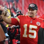 Ein Footballspieler im roten Trikot mit der Nummer 15 lächelt, während er den Fans Autogramme gibt und das dominante Playoff-Bild seines Teams feiert. Er trägt eine Kappe mit dem Logo der „Champions“. Umgeben von Fans in Rot und Gelb schaffen die roten Sitze des Stadions die Kulisse für diesen siegreichen Moment. Diese Beschreibung wurde mit der FootballR KI automatisch generiert.