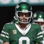 Aaron Rodgers in einer grün-weißen Uniform der New York Jets mit der Nummer 8 und einem „NY“-Logo auf dem Helm steht auf einem Feld. Auf seinem Trikot trägt er einen Kapitänsaufnäher, der an Aaron Rodgers erinnert. Der Hintergrund ist verschwommen und zeigt andere Spieler in ähnlichen Uniformen. Maxx Crosby wünscht sich den Star-QB in Las Vegas. Diese Beschreibung wurde mit der FootballR KI automatisch generiert.