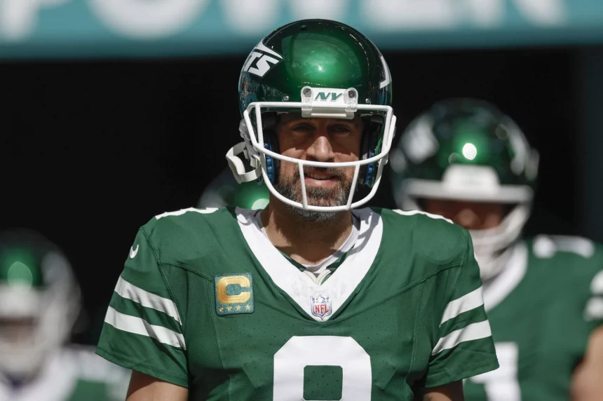Aaron Rodgers in einer grün-weißen Uniform der New York Jets mit der Nummer 8 und einem „NY“-Logo auf dem Helm steht auf einem Feld. Auf seinem Trikot trägt er einen Kapitänsaufnäher, der an Aaron Rodgers erinnert. Der Hintergrund ist verschwommen und zeigt andere Spieler in ähnlichen Uniformen. Maxx Crosby wünscht sich den Star-QB in Las Vegas. Diese Beschreibung wurde mit der FootballR KI automatisch generiert.
