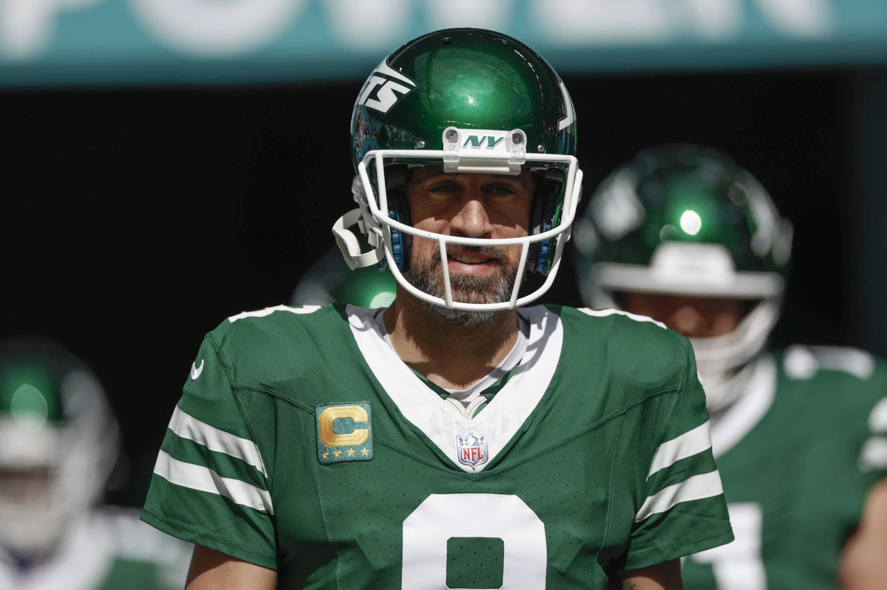 Aaron Rodgers in einer grün-weißen Uniform der New York Jets mit der Nummer 8 und einem „NY“-Logo auf dem Helm steht auf einem Feld. Auf seinem Trikot trägt er einen Kapitänsaufnäher, der an Aaron Rodgers erinnert. Der Hintergrund ist verschwommen und zeigt andere Spieler in ähnlichen Uniformen. Maxx Crosby wünscht sich den Star-QB in Las Vegas. Diese Beschreibung wurde mit der FootballR KI automatisch generiert.