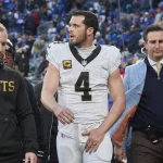 Ein Footballspieler, Quarterback Derek Carr, trägt ein weißes Trikot mit der Nummer 4 und verlässt in Begleitung von Mitarbeitern das Spielfeld. Ein Mitarbeiter trägt einen „Saints“-Kapuzenpulli, während ein anderer einen blau karierten Blazer trägt. Im Hintergrund sind eine Menge Zuschauer und eine verschwommene Stadionumgebung zu sehen. Diese Beschreibung wurde mit der FootballR KI automatisch generiert.