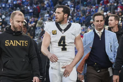 Ein Footballspieler, Quarterback Derek Carr, trägt ein weißes Trikot mit der Nummer 4 und verlässt in Begleitung von Mitarbeitern das Spielfeld. Ein Mitarbeiter trägt einen „Saints“-Kapuzenpulli, während ein anderer einen blau karierten Blazer trägt. Im Hintergrund sind eine Menge Zuschauer und eine verschwommene Stadionumgebung zu sehen. Diese Beschreibung wurde mit der FootballR KI automatisch generiert.