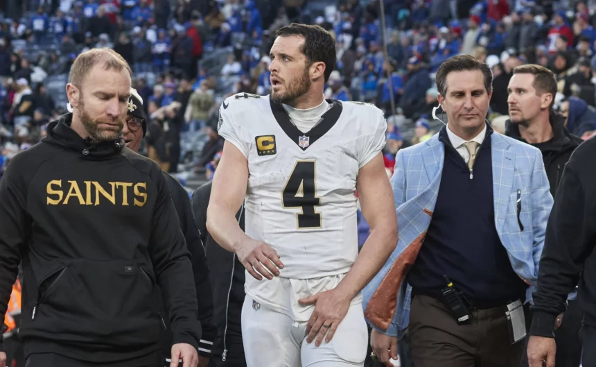 Ein Footballspieler, Quarterback Derek Carr, trägt ein weißes Trikot mit der Nummer 4 und verlässt in Begleitung von Mitarbeitern das Spielfeld. Ein Mitarbeiter trägt einen „Saints“-Kapuzenpulli, während ein anderer einen blau karierten Blazer trägt. Im Hintergrund sind eine Menge Zuschauer und eine verschwommene Stadionumgebung zu sehen. Diese Beschreibung wurde mit der FootballR KI automatisch generiert.