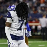 Trevon Diggs trägt ein weißes Trikot mit der Nummer 7 und lange Dreadlocks auf dem Feld. Er scheint nach unten zu blicken, während im Hintergrund Cheerleader in blau-weißen Outfits auftreten, eingerahmt von einem lebhaften Publikum und hellen Stadionlichtern. Diese Beschreibung wurde mit der FootballR KI automatisch generiert.