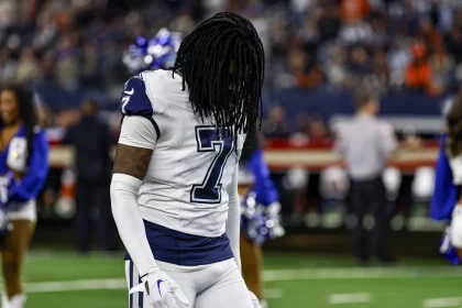 Trevon Diggs trägt ein weißes Trikot mit der Nummer 7 und lange Dreadlocks auf dem Feld. Er scheint nach unten zu blicken, während im Hintergrund Cheerleader in blau-weißen Outfits auftreten, eingerahmt von einem lebhaften Publikum und hellen Stadionlichtern. Diese Beschreibung wurde mit der FootballR KI automatisch generiert.