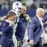 Ein Footballspieler in einer Uniform der Dallas Cowboys, identifiziert als DeMarvion Overshown, wird von zwei Mitarbeitern vom Feld geführt. Sie gehen zusammen an der Seitenlinie entlang, wobei ihn auf jeder Seite ein Mann stützt. Ein weiterer Mann im Anzug geht voran und zeigt nach vorne. Diese Beschreibung wurde mit der FootballR KI automatisch generiert.