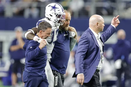 Ein Footballspieler in einer Uniform der Dallas Cowboys, identifiziert als DeMarvion Overshown, wird von zwei Mitarbeitern vom Feld geführt. Sie gehen zusammen an der Seitenlinie entlang, wobei ihn auf jeder Seite ein Mann stützt. Ein weiterer Mann im Anzug geht voran und zeigt nach vorne. Diese Beschreibung wurde mit der FootballR KI automatisch generiert.
