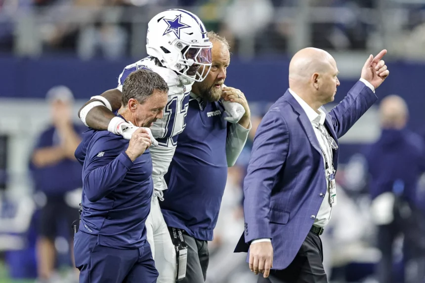 Ein Footballspieler in einer Uniform der Dallas Cowboys, identifiziert als DeMarvion Overshown, wird von zwei Mitarbeitern vom Feld geführt. Sie gehen zusammen an der Seitenlinie entlang, wobei ihn auf jeder Seite ein Mann stützt. Ein weiterer Mann im Anzug geht voran und zeigt nach vorne. Diese Beschreibung wurde mit der FootballR KI automatisch generiert.
