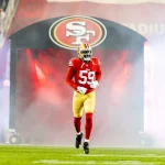 Ein Footballspieler in rot-goldener Uniform und Helm mit der Nummer 59 stürmte aus einem Tunnel mit dem Logo der San Francisco 49ers auf das Spielfeld. Rauch wirbelte um De'Vondre Campbell herum, im Hintergrund jubelten Fans und Lichter erhellten die energiegeladene Szene. Diese Beschreibung wurde mit der FootballR KI automatisch generiert.