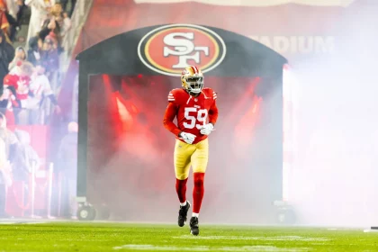 Ein Footballspieler in rot-goldener Uniform und Helm mit der Nummer 59 stürmte aus einem Tunnel mit dem Logo der San Francisco 49ers auf das Spielfeld. Rauch wirbelte um De'Vondre Campbell herum, im Hintergrund jubelten Fans und Lichter erhellten die energiegeladene Szene. Diese Beschreibung wurde mit der FootballR KI automatisch generiert.