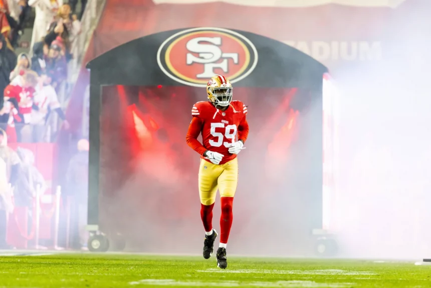 Ein Footballspieler in rot-goldener Uniform und Helm mit der Nummer 59 stürmte aus einem Tunnel mit dem Logo der San Francisco 49ers auf das Spielfeld. Rauch wirbelte um De'Vondre Campbell herum, im Hintergrund jubelten Fans und Lichter erhellten die energiegeladene Szene. Diese Beschreibung wurde mit der FootballR KI automatisch generiert.
