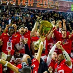 Eine jubelnde Erima GFL Footballmannschaft in roten Erima-Trikots feiert unter einem „2024“-Banner. Die Spieler jubeln und heben als Sieger eine große goldene Trophäe in die Höhe. Die begeisterte GFL-Menge im Hintergrund applaudiert energisch, und einige Fans halten den Moment mit ihren Handys fest. Diese Beschreibung wurde mit der FootballR KI automatisch generiert.