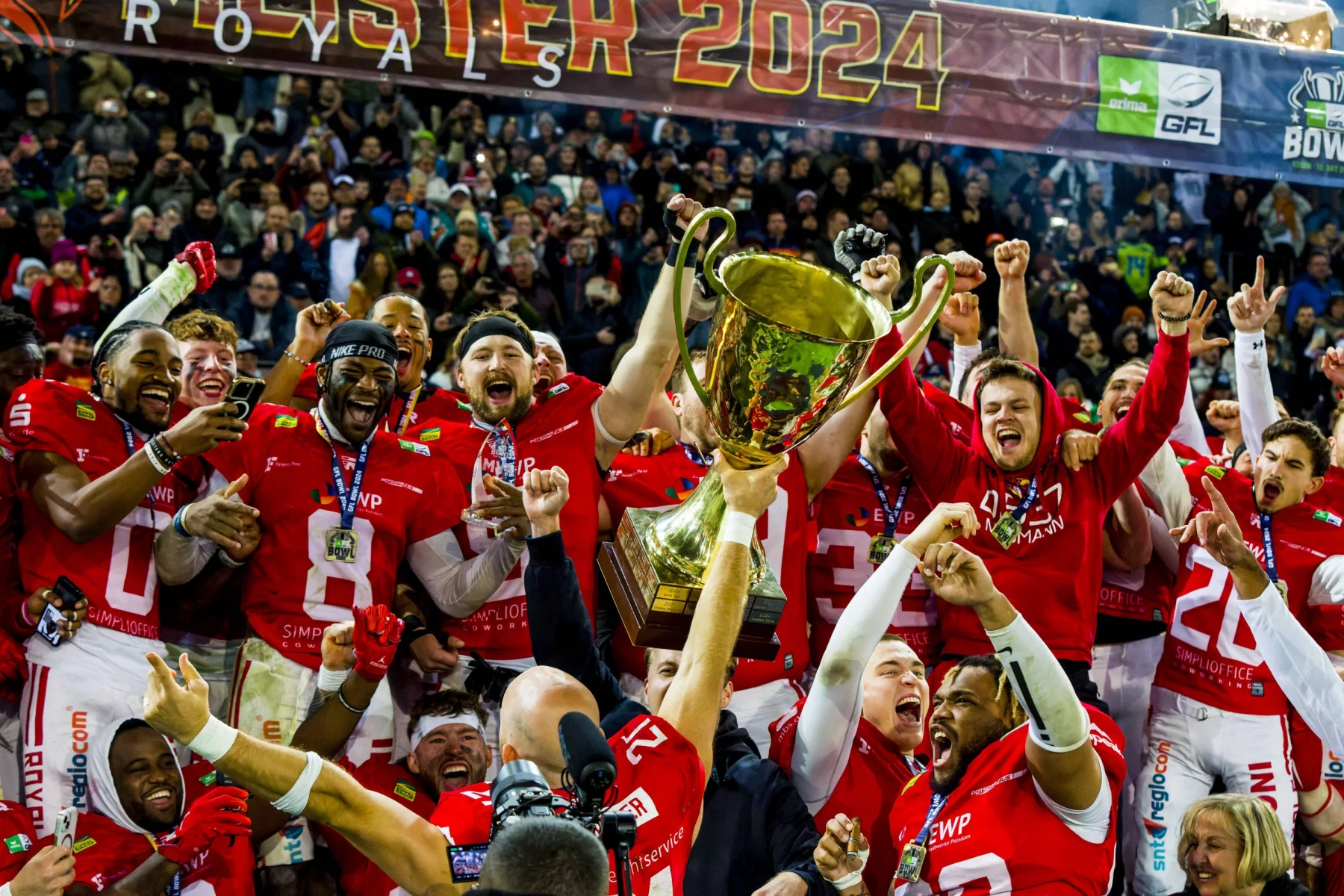 Eine jubelnde Erima GFL Footballmannschaft in roten Erima-Trikots feiert unter einem „2024“-Banner. Die Spieler jubeln und heben als Sieger eine große goldene Trophäe in die Höhe. Die begeisterte GFL-Menge im Hintergrund applaudiert energisch, und einige Fans halten den Moment mit ihren Handys fest. Diese Beschreibung wurde mit der FootballR KI automatisch generiert.
