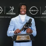 Travis Hunter steht in einem hellblauen Anzug da und hält stolz die Heisman Trophy vor einem Hintergrund aus Nissan- und Heisman-Logos in den Händen. Mit einem warmen Lächeln kombiniert er einen weißen Rollkragenpullover mit seinen langen, geflochtenen Haaren und feiert das ikonische Design der Footballspieler-Trophäe. Diese Beschreibung wurde mit der FootballR KI automatisch generiert.