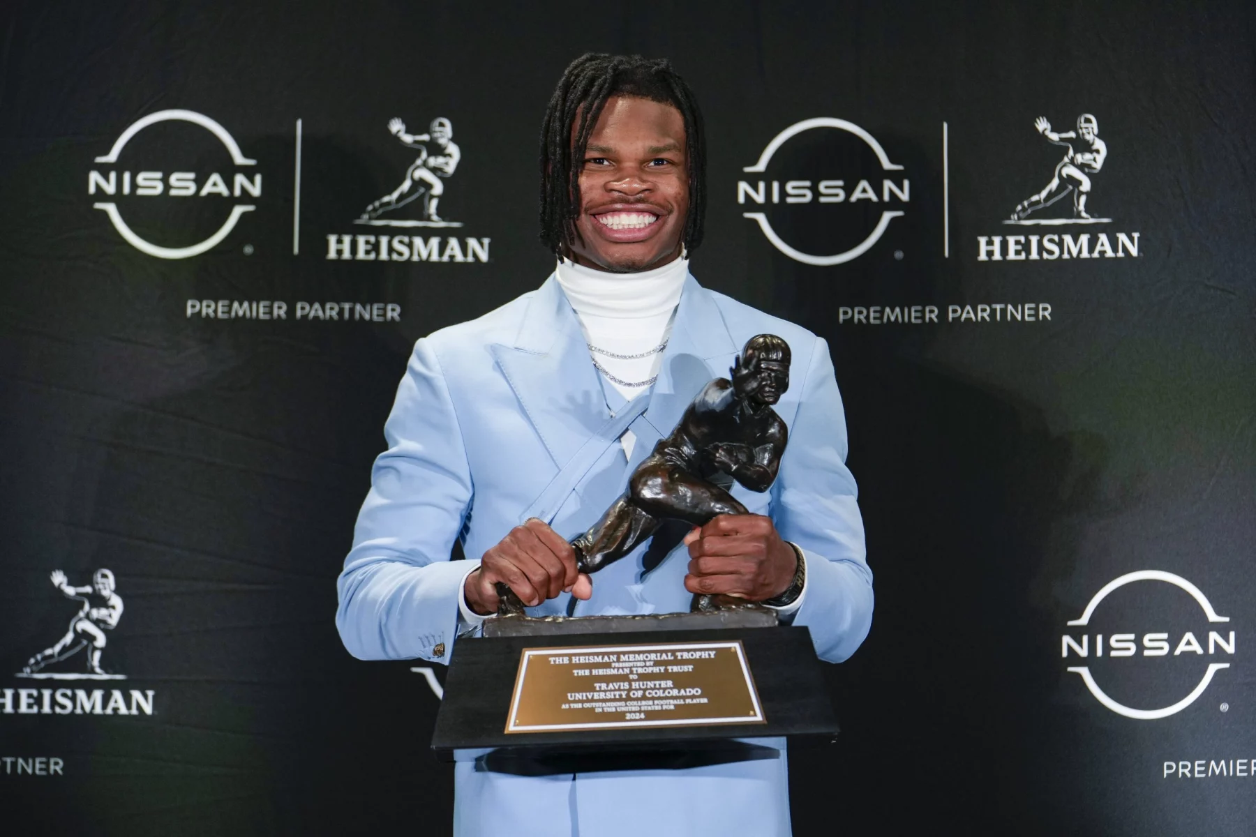 Travis Hunter steht in einem hellblauen Anzug da und hält stolz die Heisman Trophy vor einem Hintergrund aus Nissan- und Heisman-Logos in den Händen. Mit einem warmen Lächeln kombiniert er einen weißen Rollkragenpullover mit seinen langen, geflochtenen Haaren und feiert das ikonische Design der Footballspieler-Trophäe. Diese Beschreibung wurde mit der FootballR KI automatisch generiert.
