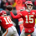 Ein Footballspieler mit der Nummer 15 in roter Uniform und Helm, der an Patrick Mahomes erinnert, bereitet sich während eines Spiels auf einen Pass vor. Ein anderer Spieler mit der Nummer 10 läuft in der Nähe. Im Hintergrund sind unscharfe Spieler und eine Menschenmenge zu sehen. Beide tragen weiße Hosen mit roten und gelben Akzenten. Diese Beschreibung wurde mit der FootballR KI automatisch generiert.