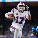 NFL MVP - NFL Parität - Hard Knocks - NFL Pro Bowl - Ein Footballspieler in weißer Uniform mit blauen und roten Akzenten, Nummer 17 Josh Allen, und rennt mit dem Ball durch ein Stadion. Er ist mitten im Schritt, hält den Ball in einer Hand und hat die andere erhoben. Im Vordergrund ist ein weiterer Spieler in schwarzer Uniform mit blauen Details zu sehen. Die Menge dahinter ist verschwommen. Diese Beschreibung wurde mit der FootballR KI automatisch generiert.