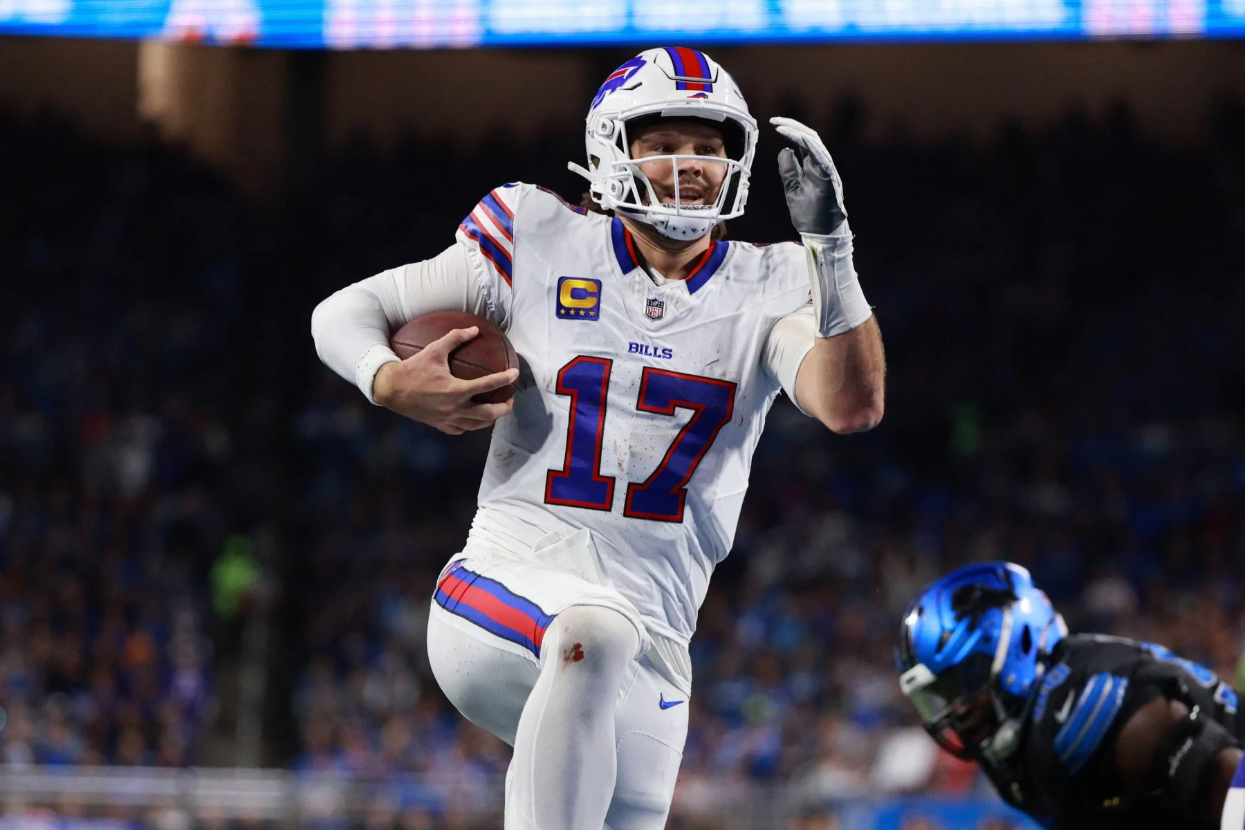 NFL MVP - NFL Parität - Hard Knocks - NFL Pro Bowl - Ein Footballspieler in weißer Uniform mit blauen und roten Akzenten, Nummer 17 Josh Allen, und rennt mit dem Ball durch ein Stadion. Er ist mitten im Schritt, hält den Ball in einer Hand und hat die andere erhoben. Im Vordergrund ist ein weiterer Spieler in schwarzer Uniform mit blauen Details zu sehen. Die Menge dahinter ist verschwommen. Diese Beschreibung wurde mit der FootballR KI automatisch generiert.