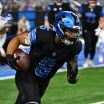 Ein Footballspieler, David Montgomery, in einer schwarzen Uniform mit blauen Akzenten, rennt mit dem Ball über das Feld. Auf seinem Helm prangt stolz die Nummer 5. Im Hintergrund liefern sich Spieler einen erbitterten Wettkampf, darunter einer in Weiß. Das Feld ist mit leuchtend blauen und weißen Linien markiert. Diese Beschreibung wurde mit der FootballR KI automatisch generiert.