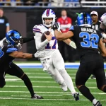 Josh Allen in weißer Uniform sprintet mit dem Ball, verfolgt von zwei Detroit Lions Spielern in schwarzen Trikots. Während ein Verteidiger nach ihm greift und ein anderer näher kommt, entfaltet sich das intensive Playoff Picture auf einem grünen Feld, wobei ein verschwommenes Publikum und andere Spieler die dramatische Szene noch verstärken. Diese Beschreibung wurde mit der FootballR KI automatisch generiert.