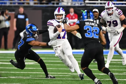 Josh Allen in weißer Uniform sprintet mit dem Ball, verfolgt von zwei Detroit Lions Spielern in schwarzen Trikots. Während ein Verteidiger nach ihm greift und ein anderer näher kommt, entfaltet sich das intensive Playoff Picture auf einem grünen Feld, wobei ein verschwommenes Publikum und andere Spieler die dramatische Szene noch verstärken. Diese Beschreibung wurde mit der FootballR KI automatisch generiert.