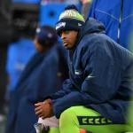 Geno Smith sitzt an der Seitenlinie und trägt eine Seattle Seahawks-Mütze und eine blaue Jacke über seiner grünen Uniform. Er wirkt konzentriert und nachdenklich, während die anderen Teammitglieder im Hintergrund verschwommen sind. Die Kulisse lässt auf einen kühlen Spieltag schließen. Diese Beschreibung wurde mit der FootballR KI automatisch generiert.