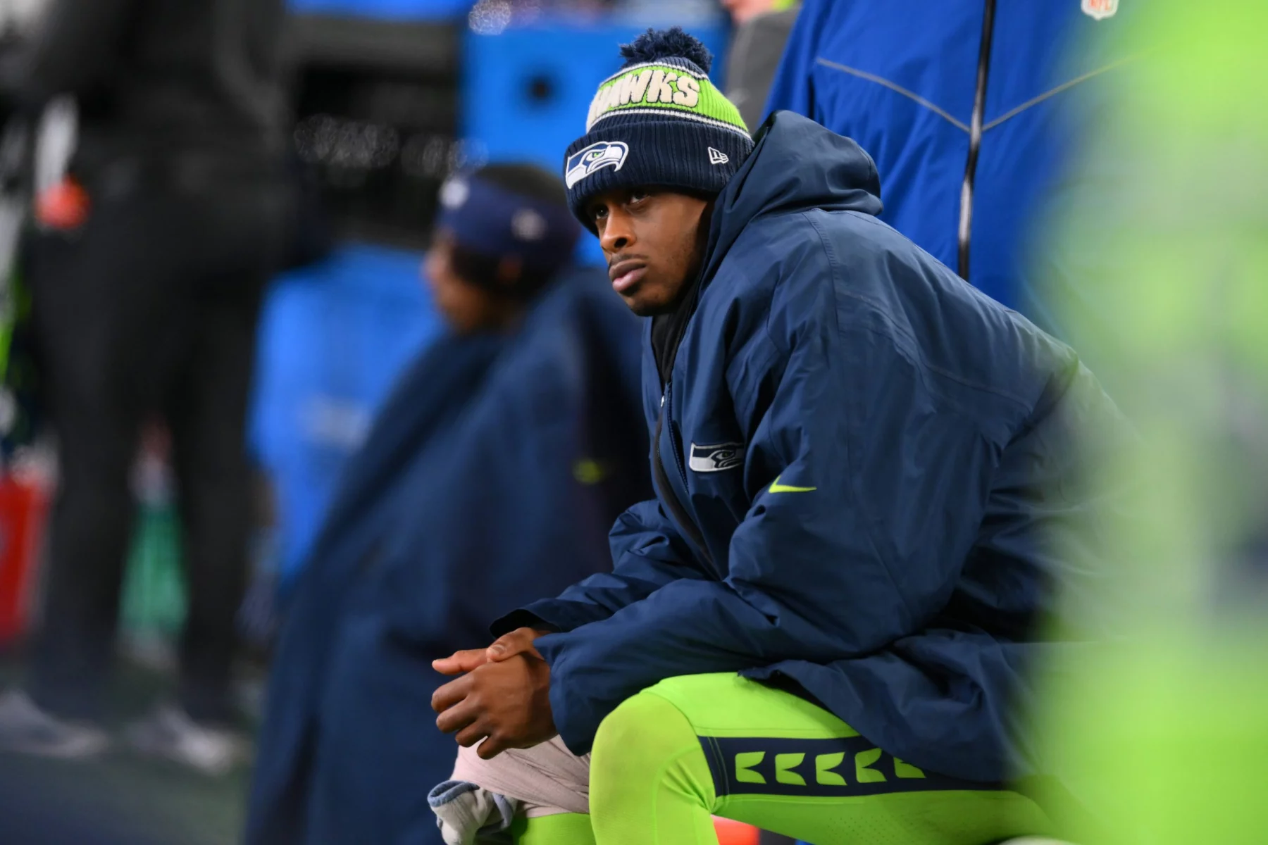 Geno Smith sitzt an der Seitenlinie und trägt eine Seattle Seahawks-Mütze und eine blaue Jacke über seiner grünen Uniform. Er wirkt konzentriert und nachdenklich, während die anderen Teammitglieder im Hintergrund verschwommen sind. Die Kulisse lässt auf einen kühlen Spieltag schließen. Diese Beschreibung wurde mit der FootballR KI automatisch generiert.