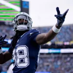 CeeDee Lamb in einer blau-silbernen Uniform gestikuliert in Richtung der Menge. Er trägt einen Helm mit Gesichtsschutz und auf seinem Trikot prangt stolz die Nummer 88. Das Stadion ist voll mit Zuschauern, doch der Gesichtsausdruck des Spielers bleibt konzentriert und entschlossen. Diese Beschreibung wurde mit der FootballR KI automatisch generiert.