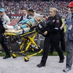 Medizinisches Personal transportiert während eines Spiels einen verletzten Footballspieler auf einer Trage, der möglicherweise in die Verletztenreserve gebracht wird. Mitarbeiter umringen Grant DuBose, der eine blaugrüne Uniform trägt. Die Szene spielt sich auf einem Feld ab, im Hintergrund ist ein volles Stadionpublikum zu sehen, während ein Mann mit einer roten Mütze zusieht. Diese Beschreibung wurde mit der FootballR KI automatisch generiert.