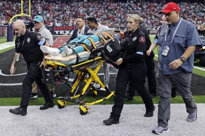 Medizinisches Personal transportiert während eines Spiels einen verletzten Footballspieler auf einer Trage, der möglicherweise in die Verletztenreserve gebracht wird. Mitarbeiter umringen Grant DuBose, der eine blaugrüne Uniform trägt. Die Szene spielt sich auf einem Feld ab, im Hintergrund ist ein volles Stadionpublikum zu sehen, während ein Mann mit einer roten Mütze zusieht. Diese Beschreibung wurde mit der FootballR KI automatisch generiert.