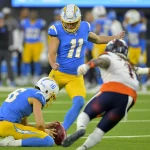 Cameron Dicker, in einem blauen Trikot und gelben Hosen, kickt ein Field Goal, während ein Teamkollege den Ball hält. Ein gegnerischer Spieler in Weiß und Orange versucht zu blocken. Das Spiel findet auf einem grünen Feld statt und fängt die Intensität jedes Augenblicks mit verschwommenen Spielern und Schiedsrichtern im Hintergrund ein. Diese Beschreibung wurde mit der FootballR KI automatisch generiert.