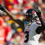 Footballspieler Tank Dell, der ein weißes Trikot der Houston Texans mit der Nummer 3 und einen Helm mit Visier trägt, streckt seine Arme nach oben auf das Spielfeld. Im Hintergrund sind verschwommene Zuschauer in Rot und Gelb sowie eine orangefarbene Seitenlinienmarkierung zu sehen. Diese Beschreibung wurde mit der FootballR KI automatisch generiert.