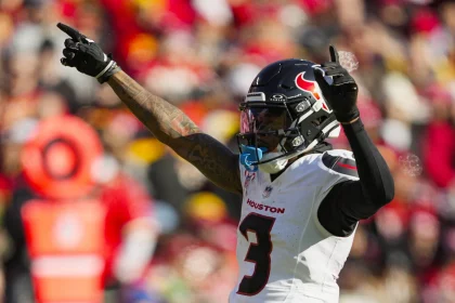 Footballspieler Tank Dell, der ein weißes Trikot der Houston Texans mit der Nummer 3 und einen Helm mit Visier trägt, streckt seine Arme nach oben auf das Spielfeld. Im Hintergrund sind verschwommene Zuschauer in Rot und Gelb sowie eine orangefarbene Seitenlinienmarkierung zu sehen. Diese Beschreibung wurde mit der FootballR KI automatisch generiert.