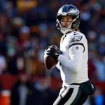 Kenny Pickett in weiß-grüner Uniform hält einen Football und ist bereit, ihn zu werfen. Mit seinem schwarzen Helm mit einem Logo an den Seiten hat er eine verblüffende Ähnlichkeit mit Kenny Pickett. Der unscharfe Hintergrund zeigt eine lebhafte Menge, die die Aufregung des laufenden Spiels einfängt. Diese Beschreibung wurde mit der FootballR KI automatisch generiert.