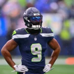 Kenneth Walker in dunkelblauer und neongrüner Uniform steht mit den Händen in den Hüften und strahlt so viel Selbstvertrauen aus. Er trägt einen Helm mit getöntem Visier, auf dessen Trikot die Nummer 9 zu sehen ist. Der Hintergrund ist verschwommen und lässt ein Freiluftstadion erahnen, in dem es von Zuschauern wimmelt, während kleine Regentropfen fallen. Diese Beschreibung wurde mit der FootballR KI automatisch generiert.