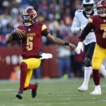 NFL Spieltag 2 - Jayden Daniels, in einer burgunderroten und goldenen Uniform, Nummer 5, läuft gekonnt mit dem Ball über das Feld. Verfolgt von Spielern wie Nummer 71, ebenfalls in ähnlichen Farben, zieht er die Zuschauer im Stadion in seinen Bann. Die Szene strahlt dynamische Sportaction unter freiem Himmel aus. Diese Beschreibung wurde mit der FootballR KI automatisch generiert.