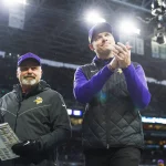 Zwei Männer auf einem Footballfeld: Kevin O'Connell, der eine schwarze Weste über einem lila Oberteil und einer Kappe trägt, applaudiert, während der andere, in einer schwarzen Jacke und einer Kappe, ein Spielblatt hält. Sie stehen am Spielfeldrand unter hellem Licht, im Hintergrund ist eine verschwommene Menschenmenge zu sehen. Diese Beschreibung wurde mit der FootballR KI automatisch generiert.