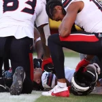 Ein Footballspieler im weißen Trikot liegt auf dem Feld, umgeben von Teamkollegen in weißen Uniformen mit schwarzen und roten Akzenten. Einer kniet neben ihm und hält den Helm des verletzten Spielers, während Tank Dell das spannende Spiel mit Sorge verfolgt. Diese Beschreibung wurde mit der FootballR KI automatisch generiert.