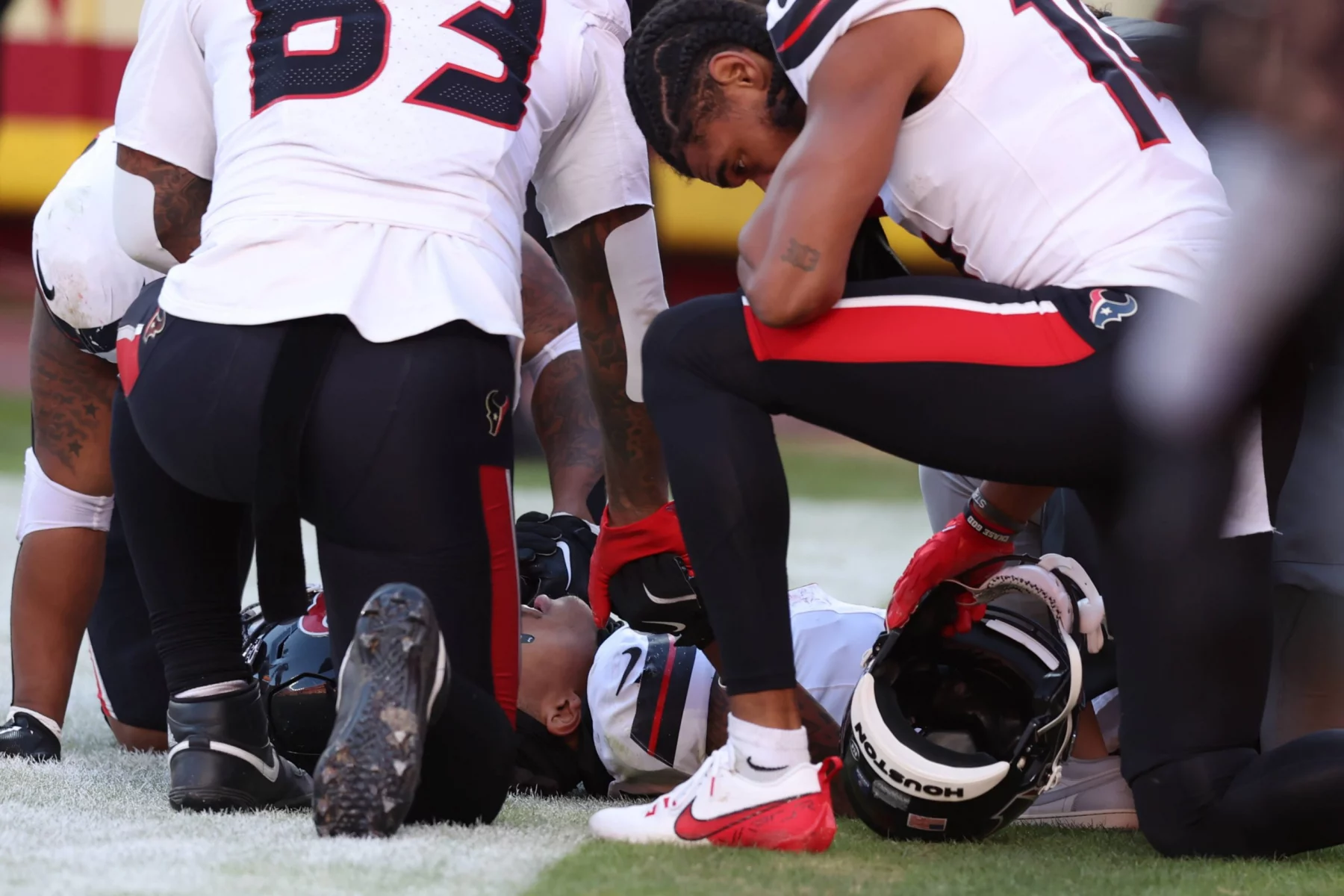 Ein Footballspieler im weißen Trikot liegt auf dem Feld, umgeben von Teamkollegen in weißen Uniformen mit schwarzen und roten Akzenten. Einer kniet neben ihm und hält den Helm des verletzten Spielers, während Tank Dell das spannende Spiel mit Sorge verfolgt. Diese Beschreibung wurde mit der FootballR KI automatisch generiert.