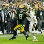 NFL TV Plan - Ein Spieler der Green Bay Packers in grün-gelber Uniform läuft mit dem Ball, dicht verfolgt von einem Spieler in weiß-schwarzer Uniform auf dem Spielfeld. Im unscharfen Hintergrund sind die Zuschauer und das Teampersonal zu sehen. Diese Beschreibung wurde mit der FootballR KI automatisch generiert.