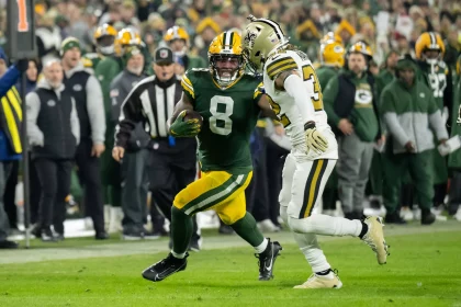 NFL TV Plan - Ein Spieler der Green Bay Packers in grün-gelber Uniform läuft mit dem Ball, dicht verfolgt von einem Spieler in weiß-schwarzer Uniform auf dem Spielfeld. Im unscharfen Hintergrund sind die Zuschauer und das Teampersonal zu sehen. Diese Beschreibung wurde mit der FootballR KI automatisch generiert.