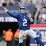 Drew Lock in einer blauen Uniform mit der Nummer 2, die an die Farben der New York Giants erinnert, wirft einen Pass auf das Spielfeld. Die dynamische Szene fängt seinen ausgestreckten Arm ein, während der Ball durch die Luft segelt, vor einem Hintergrund aus verschwommenen Zuschauern und Stadionbeschilderung. Diese Beschreibung wurde mit der FootballR KI automatisch generiert.